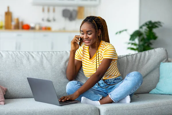 Занятая черная женщина фрилансер разговаривает по телефону и с помощью ноутбука — стоковое фото