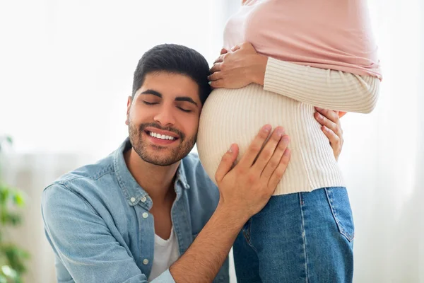 Verrukte oosterse man omhelst buik van zijn zwangere vrouw, luisterende baby kicks, teder aanraken buik van zijn echtgenoot — Stockfoto