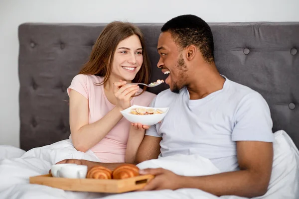 Jeune femme caucasienne nourrir son mari noir avec des céréales savoureuses dans le lit à la maison — Photo