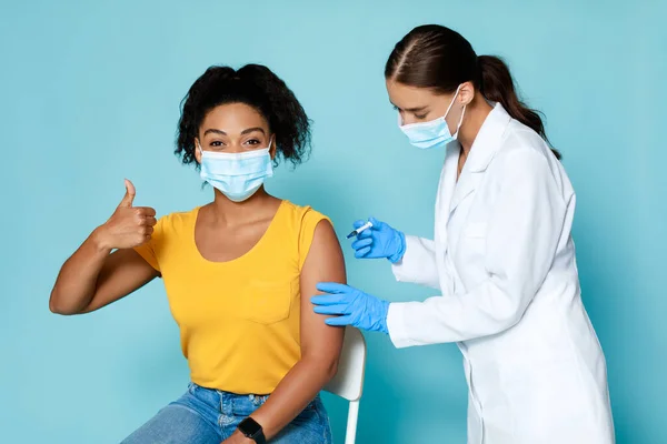 Médico femenino que administra la vacuna Covid-19 inyectable a una joven mujer negra sobre fondo azul. Vacunación poblacional —  Fotos de Stock