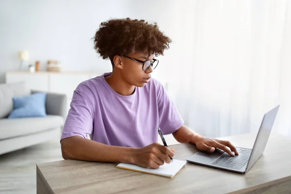 Concepto de aprendizaje electrónico. Adolescente afroamericano inteligente escribiendo en cuaderno durante la lección web en casa —  Fotos de Stock