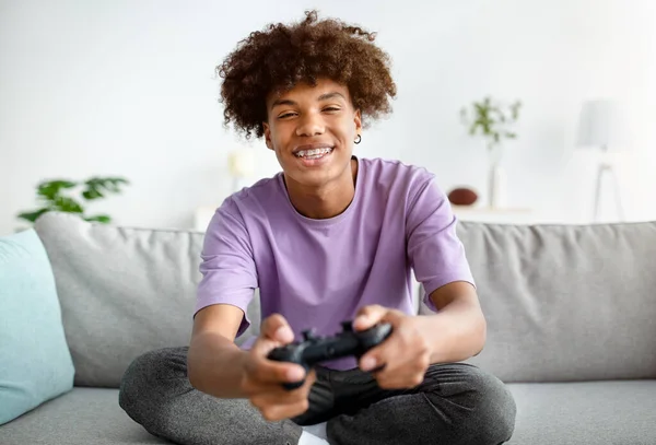 Divirtiéndose en casa. alegre negro adolescente chico con joystick jugando en línea juegos de ordenador, sentado en sofá en el interior — Foto de Stock