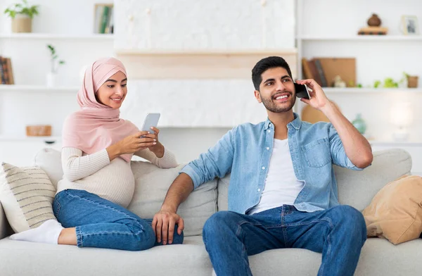 Concetto di gravidanza e tecnologia. Giovane marito musulmano e sua moglie incinta a riposo con smartphone a casa — Foto Stock