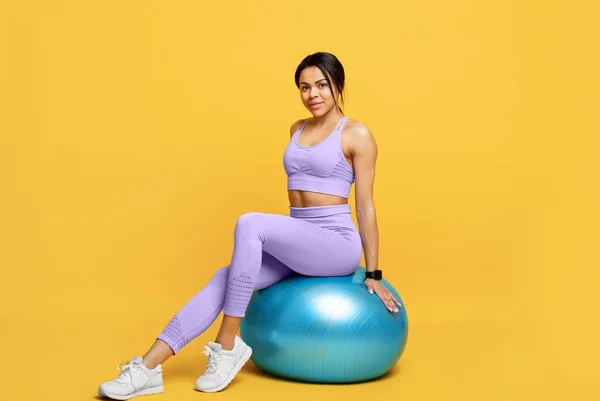 Fitness workout koncept. Glad smal afrikansk amerikansk dam sitter på fitball och ler över gul bakgrund — Stockfoto