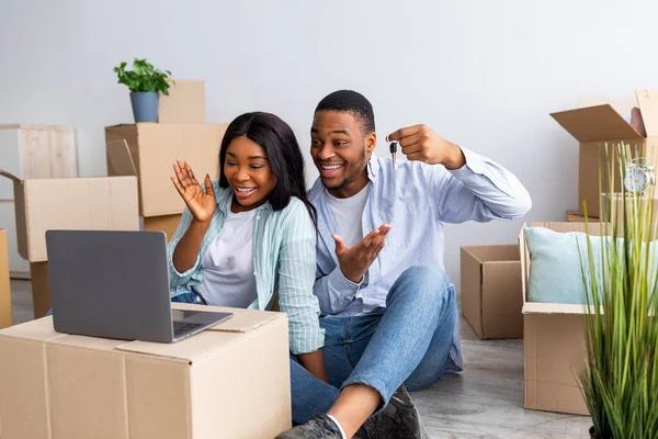 Excited black couple chatting online with friends and showing key of their house to camera, sitting among carton boxes — Stock Photo, Image