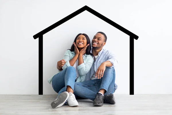 Felizes cônjuges afro-americanos sentados perto da parede branca com casa desenhada, sonhando e imaginando sua nova casa — Fotografia de Stock