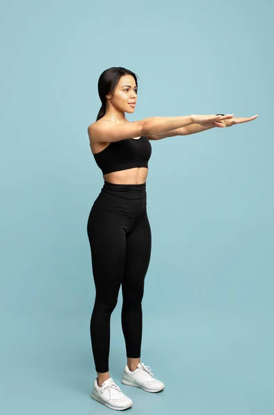 Pełna długość portret dopasowany czarny lady w sportowe ubrania robi przysiady, ćwiczenia na niebieskim tle studio — Zdjęcie stockowe