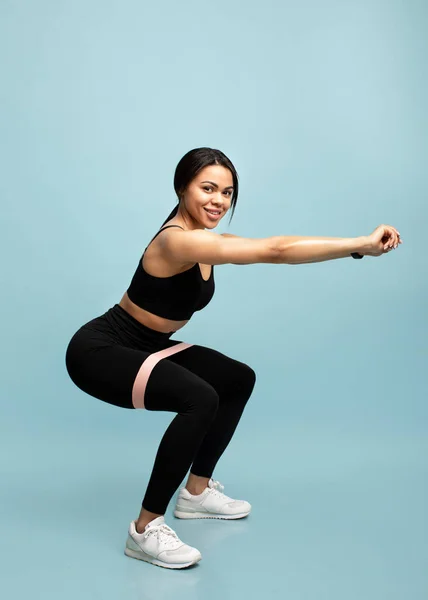 Sterke workout concept. Volledige lengte van zwarte vrouw doet squats met fitness weerstand band over blauwe achtergrond — Stockfoto