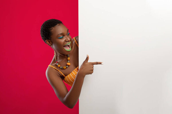 Emotional afro american short-haired young woman in stylsih summer outfit pointing at big white empty board, showing amazing advertisement or text on blank placard, red studio background