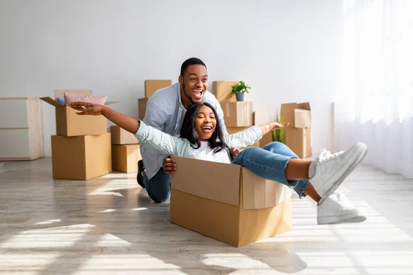 Movendo e conceito imobiliário. Joyful casal afro-americano se divertindo e montando em caixas de papelão no novo apartamento — Fotografia de Stock