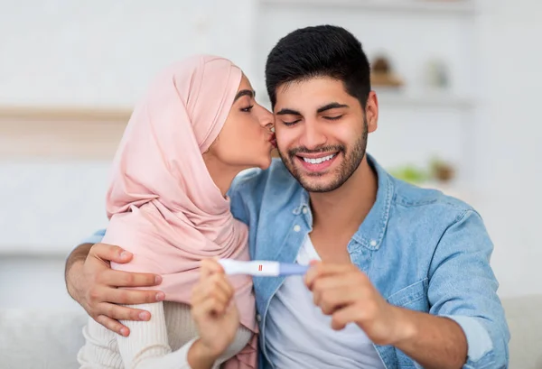 Glückliche Araberin küsst ihren Mann mit positivem Schwangerschaftstest zu Hause im Wohnzimmer — Stockfoto