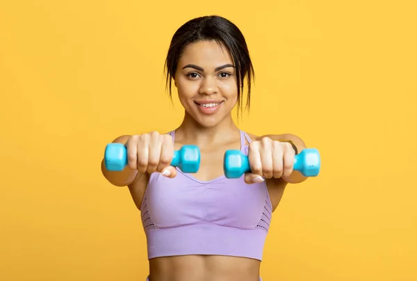 Conceito de estilo de vida saudável. Sorrindo afro-americana senhora em esportes sutiã exercitando com halteres sobre fundo amarelo — Fotografia de Stock