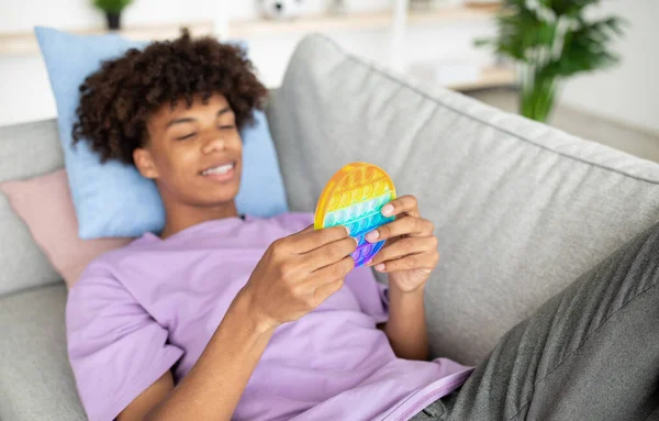 Cool black teen guy playing with bright POP IT toy, enjoying fun antistress game at home, selective focus