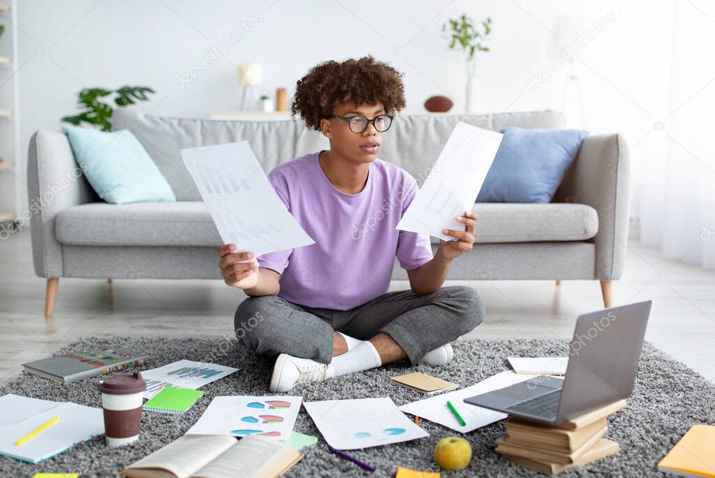 Focused black teen sitting on floor among scattered documents, using laptop to write coursework paper, studying remotely