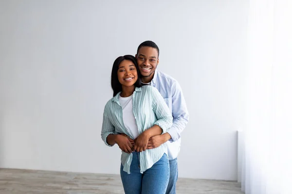 Il concetto di casa propria. Felici sposi neri che abbracciano e sorridono alla telecamera, in piedi nella stanza vuota della loro nuova casa — Foto Stock