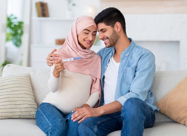 Családi és kapcsolati koncepció. Szerető terhes muszlim pár terhességi teszt otthon, ölelés és mosolygós — Stock Fotó
