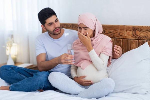 照料和支助概念。阿拉伯预产期的妻子感到不舒服，丈夫给她一杯水，坐在床上 — 图库照片