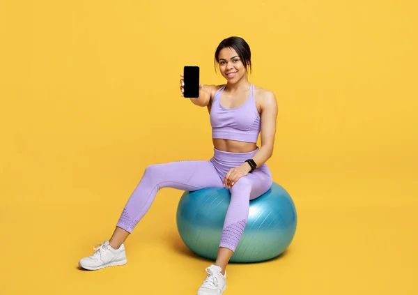 Deportiva mujer afroamericana en ropa deportiva mostrando teléfono inteligente con pantalla vacía para maqueta, sentado en fitball —  Fotos de Stock