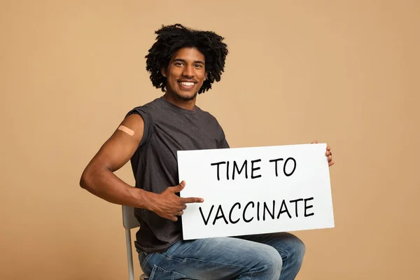 Happy black guy with time to vaccinate placard sitting over beige background — Stock Photo, Image