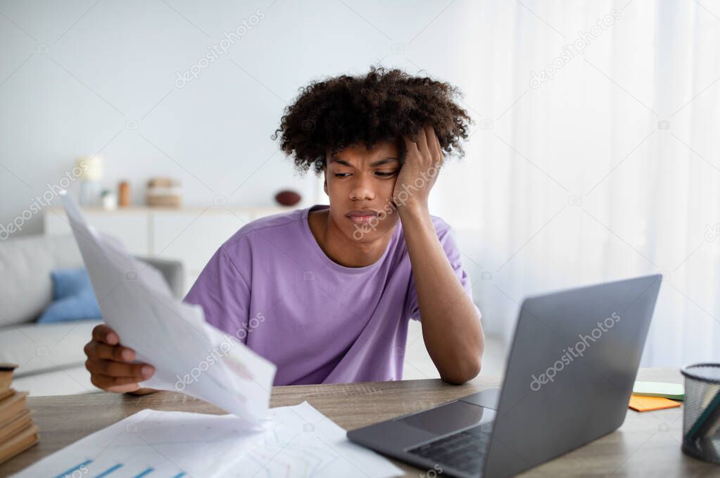 Bored teen sitting at desk with documents, using laptop to write coursework paper, studying online, preparing for exam