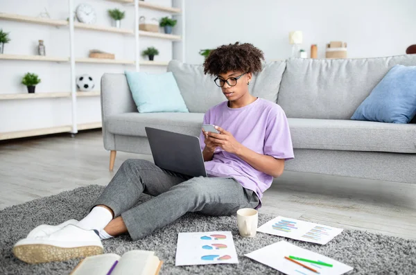 Comprimento total de focado preto adolescente cara usando smartphone e laptop para aprendizagem on-line em casa — Fotografia de Stock