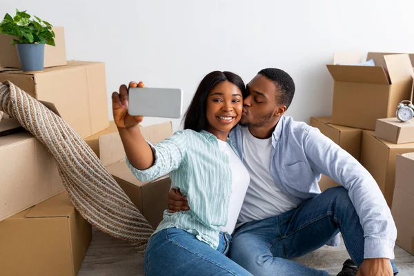Szczęśliwi czarni małżonkowie robiący selfie w swoim nowym mieszkaniu, świętujący dzień przeprowadzki, siedzący w pobliżu kartonów — Zdjęcie stockowe