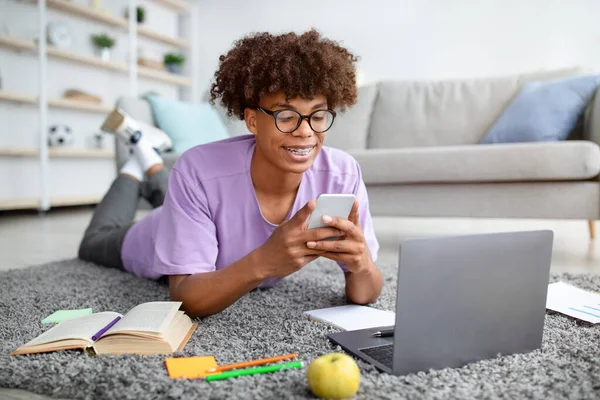 Feliz adolescente negro deitado no chão com smartphone e laptop, estudando online, verificando as mídias sociais em casa — Fotografia de Stock