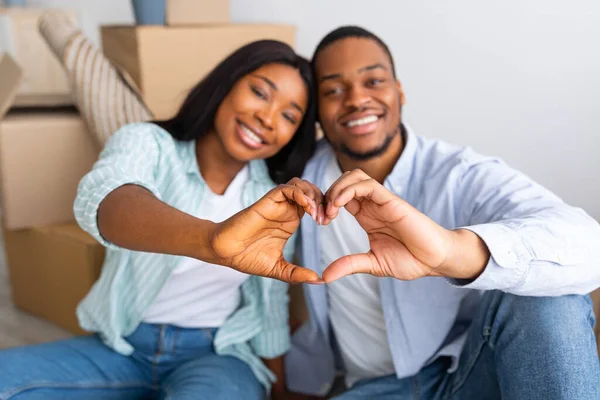 Housing for young family. Romantic black couple gesturing heart shape with fingers, sitting in new house among boxes — Stock Photo, Image