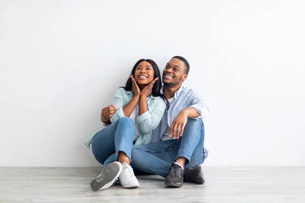 Dreaming about future. Excited black couple sitting on floor leaning on wall in new apartment, looking at free space — Stock Photo, Image