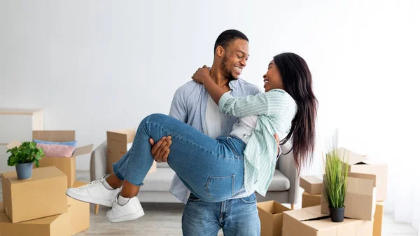 Happy moving day. Loving african american couple celebrating relocation, husband lifting his wife up on hands, panorama — Stock Photo, Image