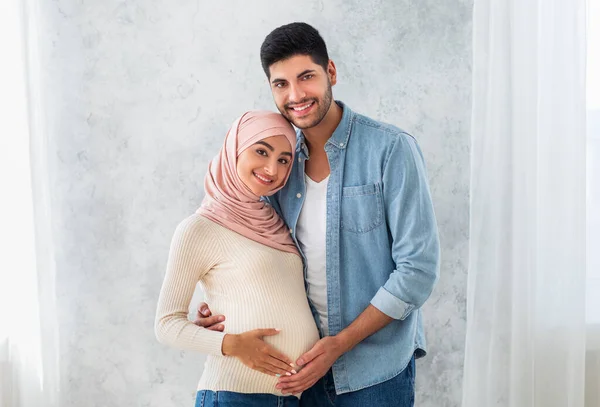 Portret van jonge zwangere Arabische echtgenoten, omhelzend en lachend naar de camera, liefhebbende moslimman knuffelend met zijn vrouw buik — Stockfoto