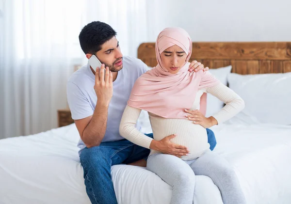 Islámica esperando madre en hiyab sufriendo espasmos dolorosos y trabajo de parto espontáneo mientras marido preocupado llamando de emergencia — Foto de Stock