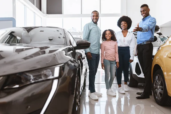 Купівля або оренда автомобіля. Радісна чорна сім'я розмовляє зі усміхненим продавцем про покупку нового автомобіля в автосалоні — стокове фото
