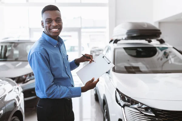 Vrolijke zwarte auto verkoper met lege klembord, glimlach op de camera in de moderne auto-dealer, mockup — Stockfoto