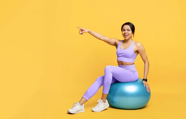 Felice in forma afro-americana signora seduta su fitball e che indica lo spazio libero su sfondo studio giallo, panorama — Foto Stock