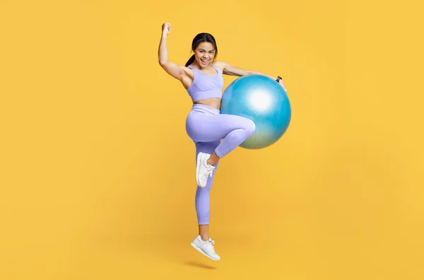 Concept de mode de vie actif. Femme noire ravie de sauter avec une balle de fitness et de célébrer le succès sur fond jaune — Photo