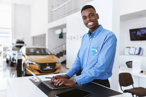 Alegre vendedor de carro preto posando na mesa de trabalho, sorrindo para a câmera, digitando no laptop em concessionária de automóveis — Fotografia de Stock