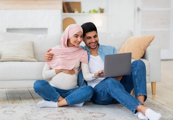 Milující těhotné arabky manželé pomocí notebooku, sedí na podlaze v obývacím pokoji, sledování interiéru dětského pokoje, volné místo — Stock fotografie