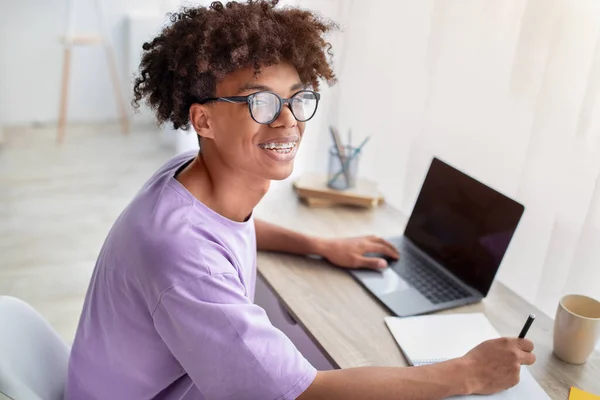 Szczęśliwy czarny nastolatek siedzi przy biurku z laptopem, uczy się zdalnie, notuje w copybooku w domu — Zdjęcie stockowe