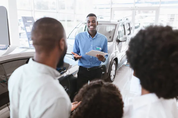 Jovem família negra escolhendo carro novo na concessionária de automóveis, vendedor ajudando-os a tomar a decisão certa — Fotografia de Stock