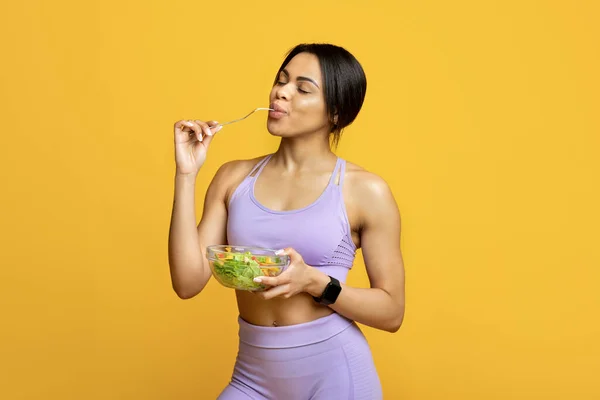 Saludable señora afroamericana deportiva disfrutando de ensalada de verduras frescas, de pie sobre fondo de estudio amarillo — Foto de Stock