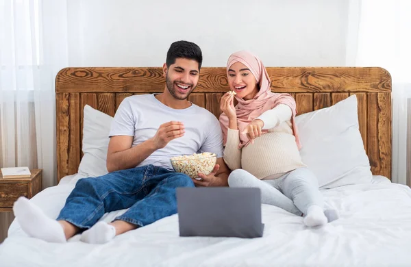 Una comedia graciosa. Hombre musulmán feliz y esposa embarazada viendo la película en el ordenador portátil, comiendo palomitas de maíz y riendo —  Fotos de Stock