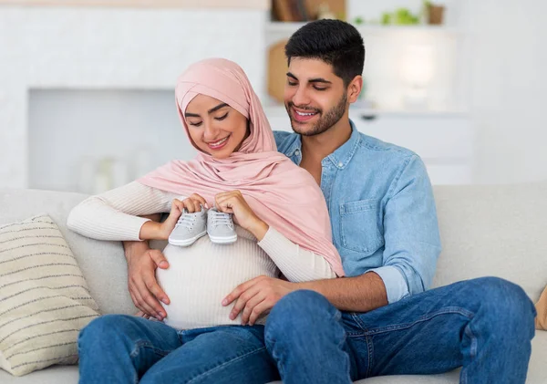 Feliz pareja musulmana embarazada colocando zapatos de bebé en el vientre grande de la mujer mientras descansan juntos en el sofá en casa — Foto de Stock