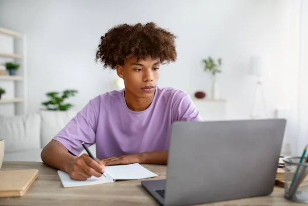 Estudiando desde el concepto del hogar. Chico adolescente negro serio tomando notas mientras participa en la lección en línea en el ordenador portátil —  Fotos de Stock