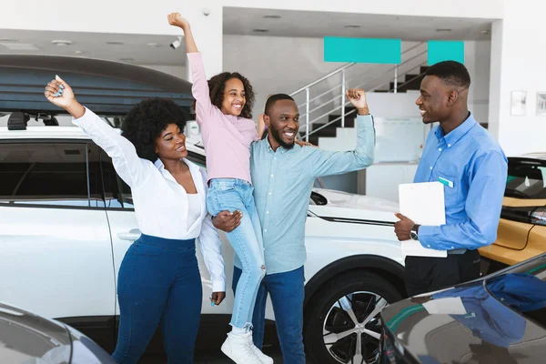 Família negra sentindo-se animado sobre a compra de automóveis, levantando as mãos, gestos SIM, de pé com vendedor de carros na concessionária — Fotografia de Stock