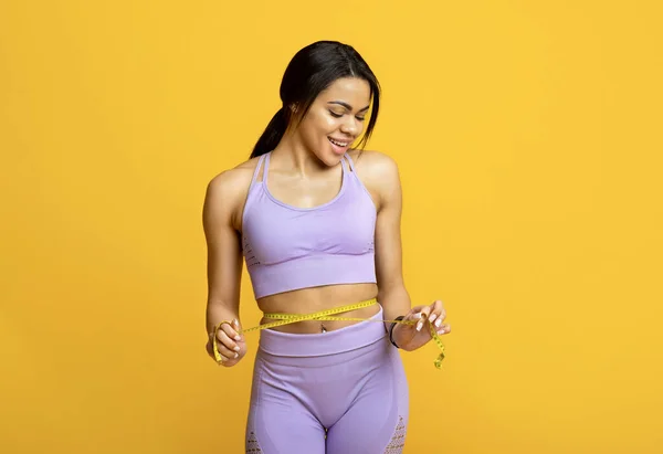 Happy fit young black woman measuring waist with yellow tape and smiling, isolated on yellow background — Stock Photo, Image