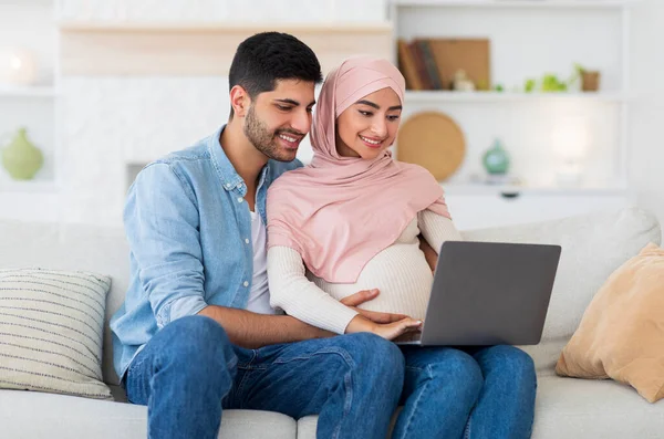 Szerető terhes muszlim pár laptoppal, pihenés kanapén a nappaliban, együtt töltött idő, üres tér — Stock Fotó