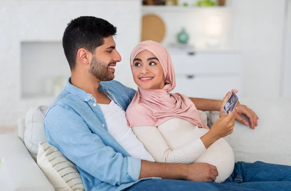 Esposos islâmicos felizes olhando para a foto de ultra-som de seu futuro bebê descansando em casa no sofá, espaço livre — Fotografia de Stock