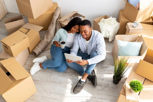 Familia afroamericana feliz sentada en el piso en nuevo apartamento con tableta entre cajas con pertenencias, arriba ver — Foto de Stock