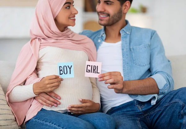 Género revelar concepto. Feliz pareja árabe sosteniendo tarjetas adhesivas de niño y niña en colores rosa y azul, sentado en el sofá — Foto de Stock